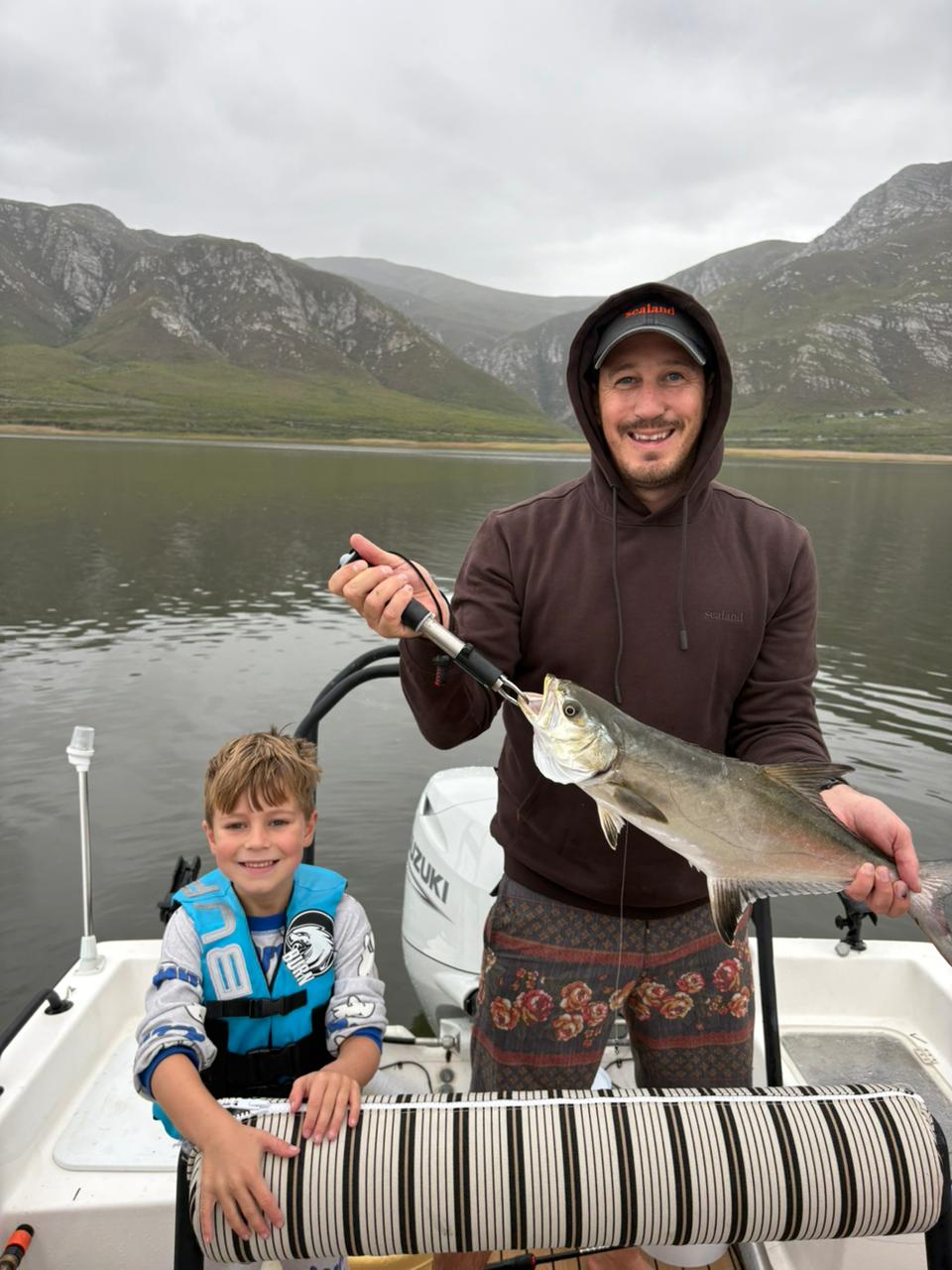 Father and son on a boat holding a large fish with mountains behind